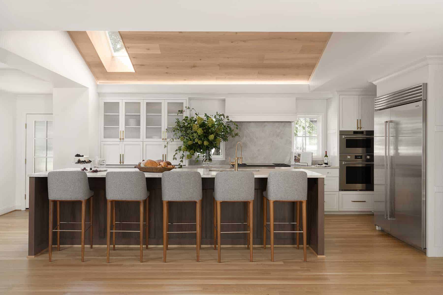 Installed by Central Surfaces, Taj Mahal quartzite in a leathered finish wraps the backsplash wall and matching countertops. White oak engineered wide-plank flooring brightens the space, while a skylight above the vaulted ceiling filters natural light into the kitchen.